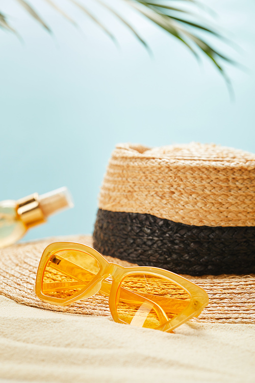 selective focus of sunglasses near straw hat and bottle with suntan oil on sandy beach isolated on blue