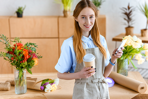 beautiful young florist holding smartphone and coffee to go, smiling at camera in flower shop