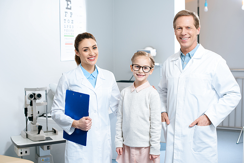 two smiling ophthalmologists and little child in glasses standing in clinic