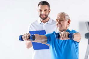 portrait of rehabilitation therapist with notepad assisting senior man exercising with dumbbells on grey backdrop
