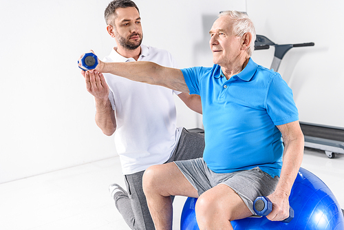 rehabilitation therapist assisting senior man exercising with dumbbells on fitness ball
