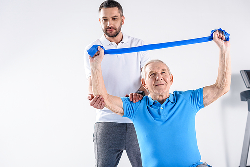 portrait of rehabilitation therapist assisting senior man exercising with rubber tape on grey backdrop