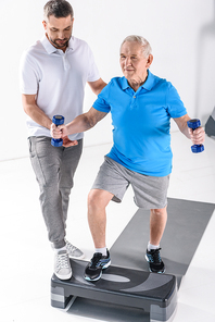 rehabilitation therapist assisting senior man exercising with dumbbells on stepper on grey backdrop