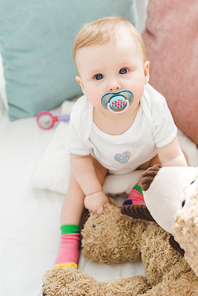 High angle view of infant with baby dummy and teddy bear sitting in crib