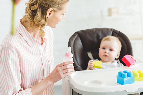 Young mother holding feeding bottle and infant daughter sitting in baby chair with plastic blocks