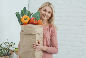 portrait of smiling mature woman with paper bag full of fresh vegetables in hands at home