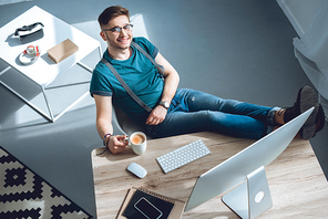 high angle view of handsome young man in eyeglasses smiling at camera while sitting at workplace at home office