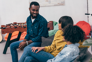 group of multicultural smiling friends sitting on couch and talking to each other