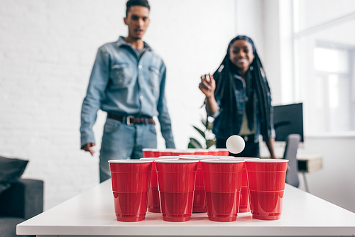 closeup shot of ball flying in plastic cup in beer pong game and young multicultural couple standing behind