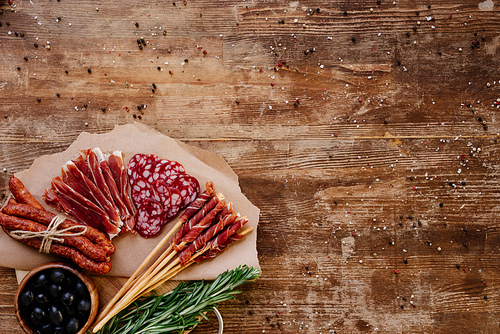 top view of round cutting board with tasty prosciutto, salami, smoked sausages, olives and herbs on wooden vintage table with scattered spices