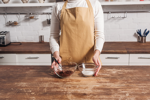 cropped shot of woman in apron holding glass bowls with ingredients for delicious homemade cake