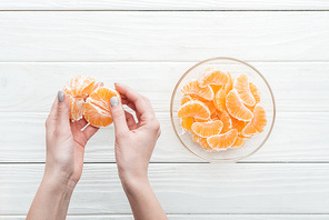 cropped view of woman holding peeled tangerine near glass bowl with tangerine slices on wooden white background
