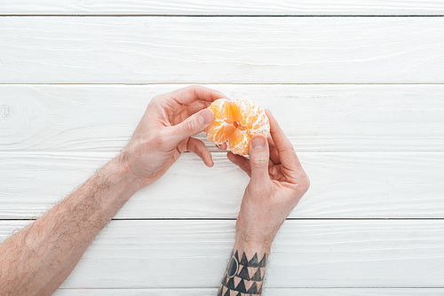 cropped view of tattooed man holding peeled tangerine on white wooden surface