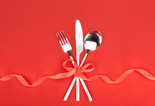 top view of  fork, knife and spoon wrapped by red festive ribbon isolated on red, st valentine day concept