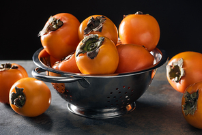 orange and ripe persimmons in colander isolated on black
