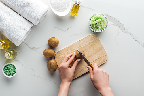 partial view of woman cutting kiwi on wooden cutting desk, and various natural ingredient for handmade cosmetics on white surface