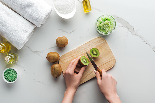 partial view of woman cutting kiwi on wooden cutting desk, and different ingredients for making cosmetics on white surface