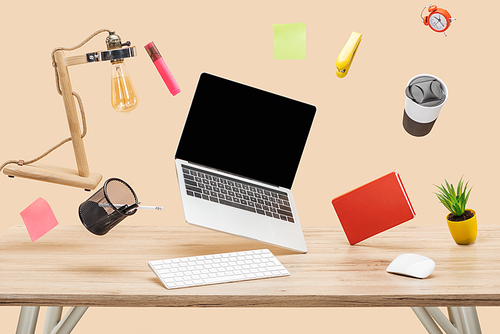 laptop with blank screen, lamp, sticky notes and stationery levitating in air above wooden desk isolated on beige