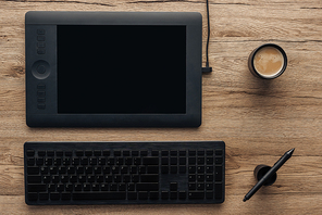 top view of black graphic tablet, graphic pen, wireless computer keyboard and cup with coffee on wooden table