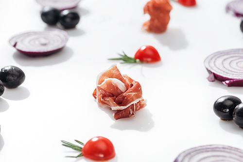 selective focus of tasty prosciutto near cherry tomatoes with rosemary twigs near red onion rings and black olives on white background