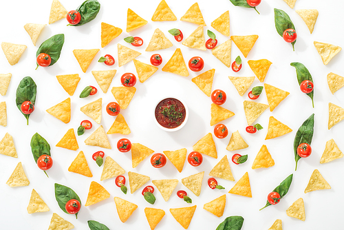 flat lay with tasty nachos, sauce and sliced chili peppers with basil and cherry tomatoes on white background