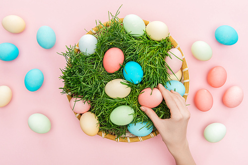 top view of female hand with easter eggs in wicker plate with grass isolated on pink