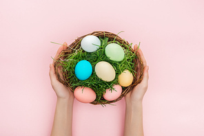 cropped view of female hands holding wicker plate with grass and easter eggs isolated on pink