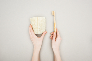 cropped view of woman holding organic sponge and bamboo toothbrush on grey background