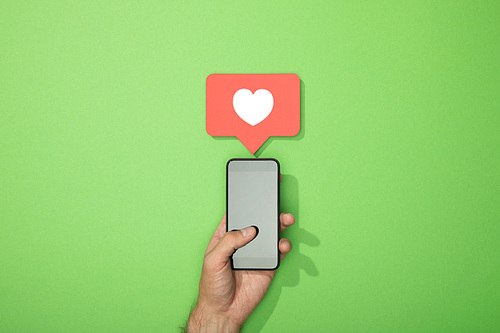 cropped view of man holding smartphone with white heart in icon above gadget on green