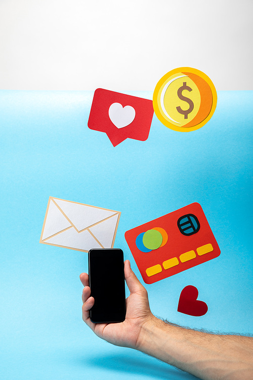 cropped view of man holding smartphone near colorful paper icons on blue and grey background