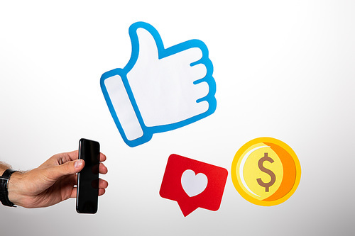 cropped view of man holding smartphone near colorful paper icons with coin, heart and thumb up on white background