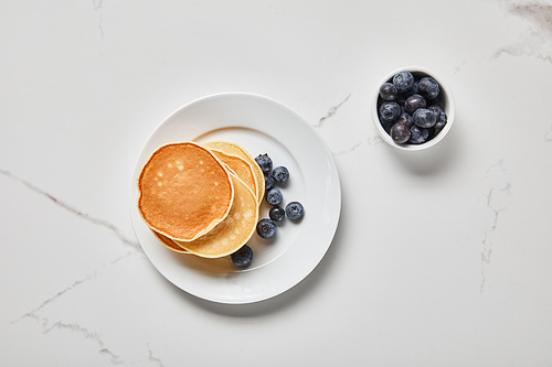 top view of pancakes in plate near bowl with blueberries on textured surface