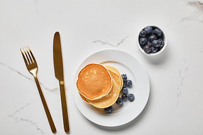 top view of pancakes on plate near golden cutlery and bowl with blueberries
