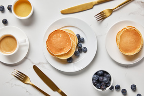 top view of pancakes, cup of coffee and scattered blueberries near golden cutlery