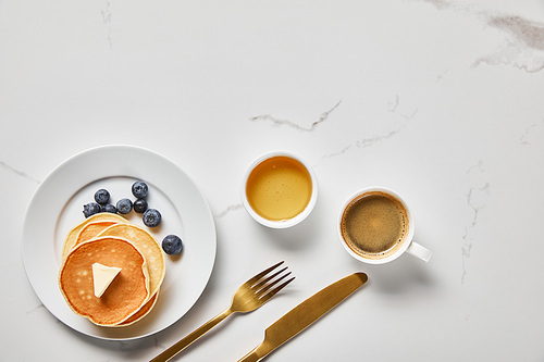 top view of bowl with honey, tasty pancakes with blueberries and cup of coffee near golden cutlery