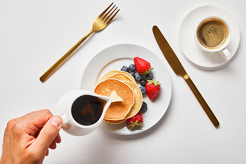 cropped view of man pouring syrup on pancakes with berries near golden cutlery and cup of coffee