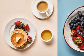 top view of pancakes with berries on plate, bowl with honey, cup of coffee and plate with berries on pink and blue background