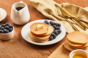 plate with pancakes and blueberries, jug with syrup near linen cloth with golden cutlery on wooden surface