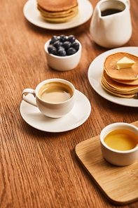 selective focus of cup of coffee on saucer near plate with pancakes and bowls with honey and blueberries on wooden surface