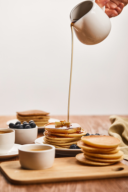 cropped view of man pouring tasty pancakes with syrup from jug near bowls with blueberries and honey, and cup of coffee isolated on grey