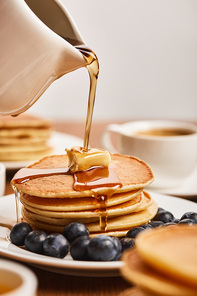 selective focus of syrup pouring on pancakes with butter and blueberries near cup of coffee