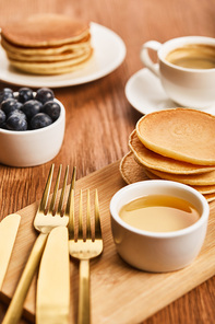 selective focus of bowl with honey, pancakes and blueberries near cup of coffee on wooden surface