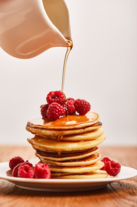 syrup pouring on pancakes with raspberries on plate on wooden surface isolated on grey