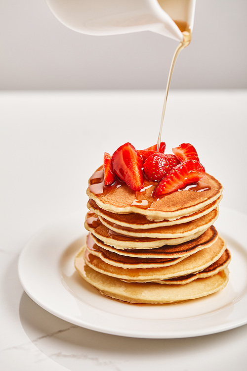 pouring syrup from jyg on tasty pancakes with strawberries on white plate on textured surface