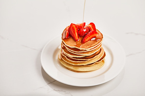 pouring syrup on tasty pancakes with strawberries on white plate on textured surface