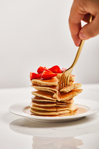 cropped view of man taking slice of pancakes with strawberries on white plate isolated on grey
