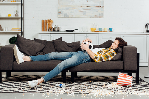 tired man holding football and sleeping on sofa near bucket and popcorn on floor