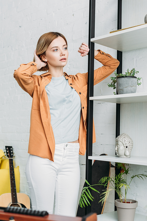 attractive girl in orange shirt and white jeans standing by shelving rack and touching hair