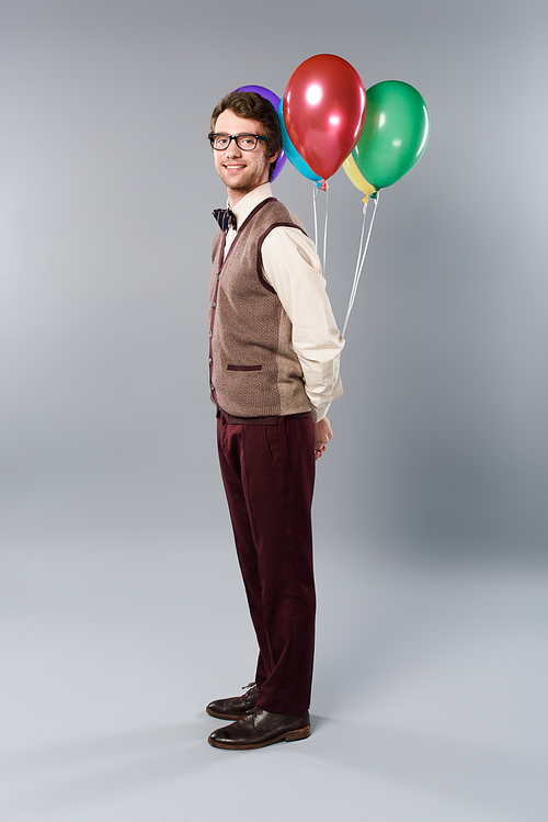 positive man in glasses holding multicolored balloons on grey background
