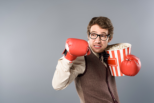 angry man in glasses and boxing gloves holding bucket of popcorn on grey background